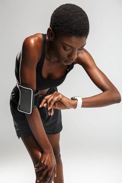 Wizerunek afrykańskiej kobiety sportowej korzystającej z telefonu komórkowego i smartwatcha podczas treningu na białym tle - Zdjęcie, obraz
