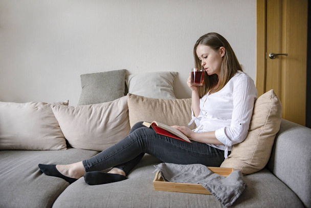 1 white girl in a white shirt sitting on a sofa in a room, drinking tea and reading a book - 写真・画像