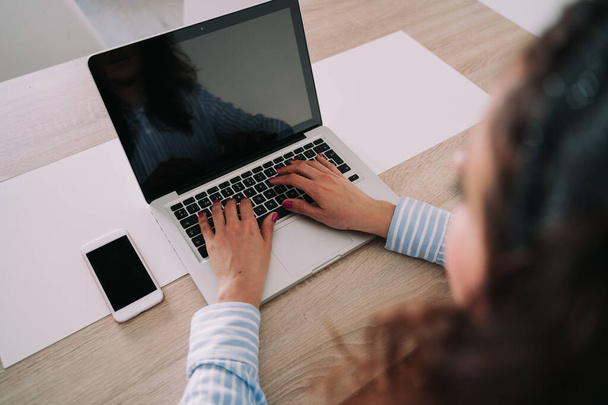 Une femme d'affaires écrit sur son ordinateur portable qui est situé sur une table en bois. Elle porte des vêtements de travail. Elle a son téléphone à côté de l'ordinateur portable
. - Photo, image