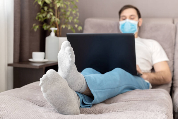 Young caucasian man working from home wearing protective mask using laptop and internet. Cozy Home office, workplace on sofa during coronavirus pandemic, covid 19 quarantine. Remote work, freelancer. - Zdjęcie, obraz