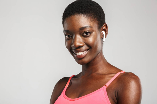 Bild einer afrikanisch-amerikanischen Sportlerin, die drahtlose Ohrhörer benutzt und isoliert auf weißem Hintergrund lächelt - Foto, Bild