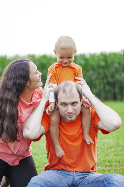 Portrait of happy family playing with son outdoors. Couple sitting together on grass, little boy sitting on neck of his father. Family, parenthood, relationships - Photo, Image