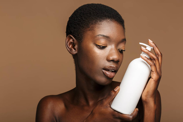 Beauty portrait of pretty young half-naked african woman holding body lotion bottle isolated over beige background - Foto, Imagen