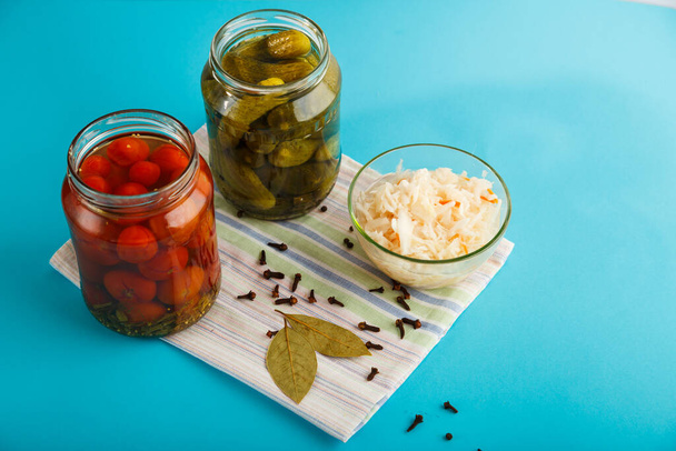 Jars with pickled cucumbers and tomatoes and sauerkraut on a wooden table on a blue background on a linen towel. - Foto, imagen