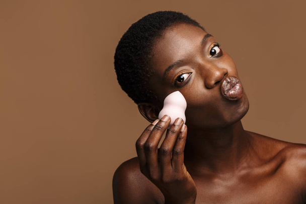 Beauty portrait of pretty young half-naked african woman holding makeup puff isolated over brown background - Zdjęcie, obraz
