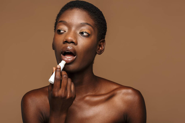 Beauty portrait of pretty young half-naked african woman putting on lipbalm isolated over brown background - Foto, afbeelding