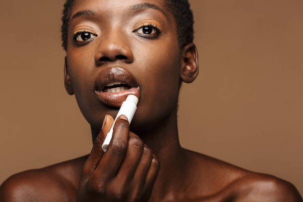 Beauty portrait of pretty young half-naked african woman putting on lipbalm isolated over brown background - Фото, зображення
