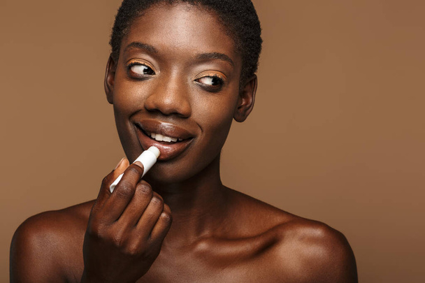 Beauty portrait of pretty young half-naked african woman putting on lipbalm isolated over brown background - Photo, image