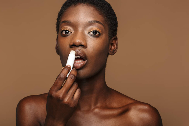 Beauty portrait of pretty young half-naked african woman putting on lipbalm isolated over brown background - Foto, afbeelding