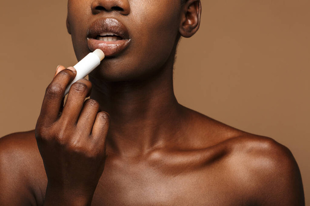 Beauty portrait of pretty young half-naked african woman putting on lipbalm isolated over brown background - Foto, Imagen