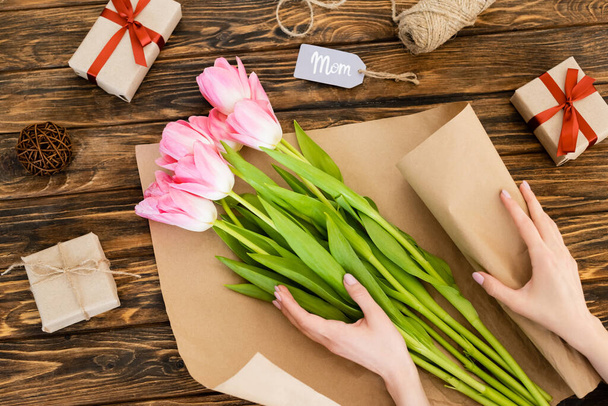 cropped view of woman wrapping pink tulips in paper near gift boxes on wooden surface, mothers day concept  - Zdjęcie, obraz
