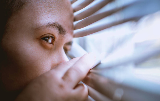 Ein Mann schaut während der Coronavirus-Pandemie zu Hause durch die Jalousien aus dem Fenster - Foto, Bild