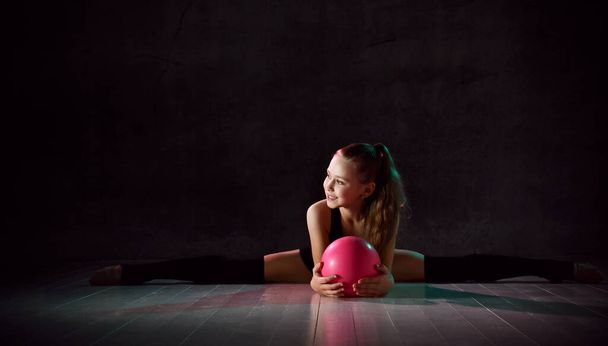 Young smiling girl gymnast in black sport body and uppers sitting in twine on floor and holding pink gymnastic ball in hands - Foto, Bild