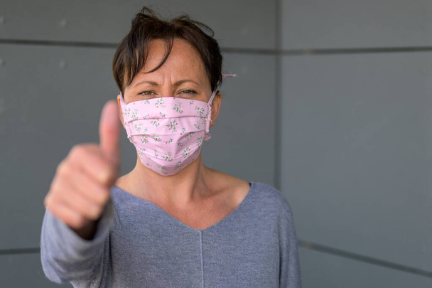 Woman wearing a face mask giving a thumbs up to show that all is well during the coronavirus or Covid-19 pandemic in a head and shoulders view - Photo, Image