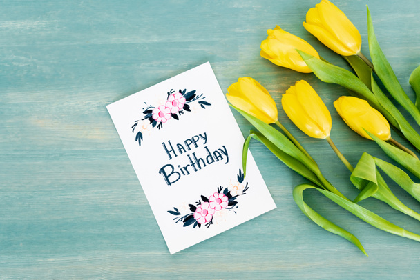 Draufsicht auf Grußkarte mit Happy Birthday Schriftzug in der Nähe von gelben Tulpen auf blau strukturierter Oberfläche  - Foto, Bild