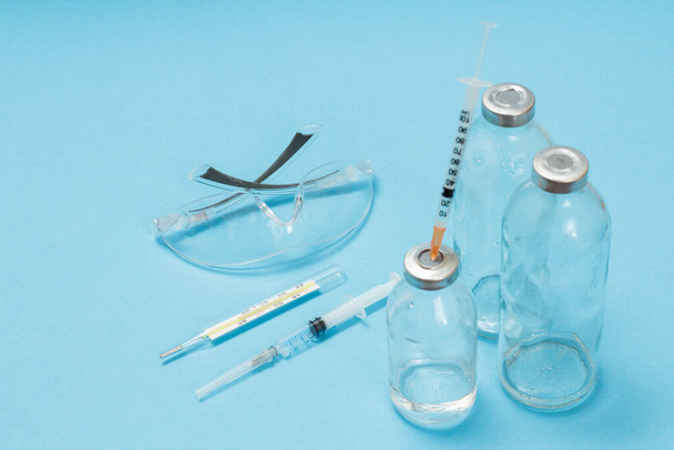 Protective glasses, glass bottles for liquid medicine, a glass mercury thermometer and syringes for injection on a blue background. Top view. Health and medical concept. - Photo, Image
