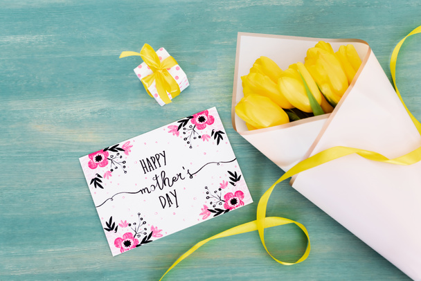 top view of small gift box near greeting card with happy mothers day lettering and yellow tulips on blue wooden surface  - Foto, immagini