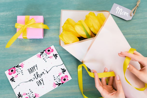 vista recortada de la mujer tocando la cinta cerca de los tulipanes amarillos, tarjeta de felicitación con letras feliz día de las madres y etiqueta de mamá en la superficie azul
  - Foto, imagen