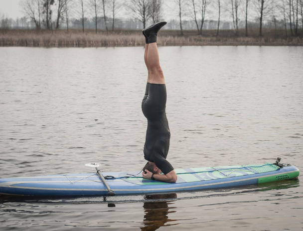 Sup Yoga, bulutlu bir günde yoga yapan dalgıç kıyafetli bir adam, kürek tahtasında amuda kalkıyor., - Fotoğraf, Görsel