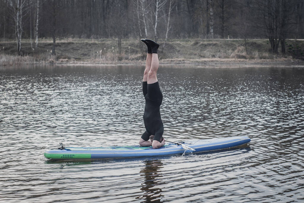 SUP YOGA, человек в гидрокостюме, занимающийся йогой в облачный день, стоя на голове на веслах
, - Фото, изображение