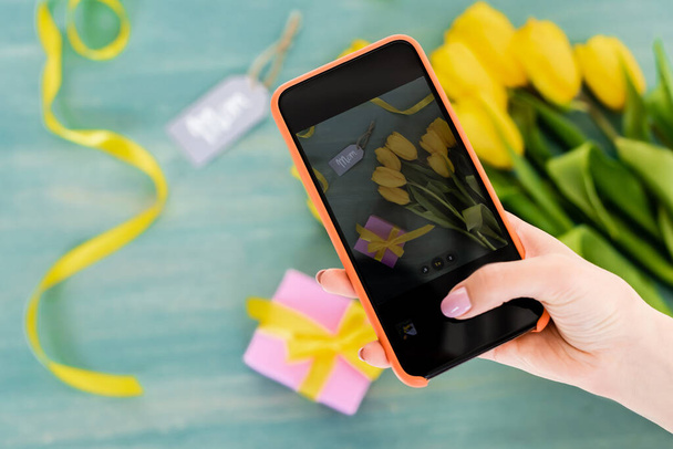 enfoque selectivo de la mujer tomando fotos de tulipanes amarillos, caja de regalo y letras de etiquetas de mamá en la superficie texturizada, concepto de día de las madres
  - Foto, Imagen