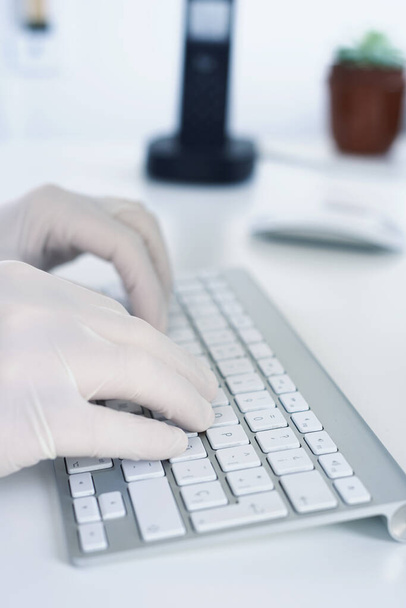 Bir adama yakın çekim, lateks eldiven takıyor, beyaz bir masada oturan bir bilgisayar klavyesine yazı yazıyor. - Fotoğraf, Görsel