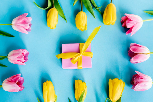 top view of blooming yellow and pink tulips near gift box on blue, mothers day concept  - 写真・画像