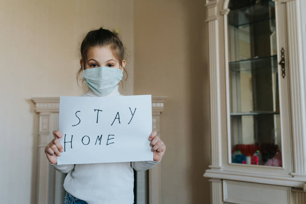 Stay at home concept. Small girl in mask holding sign saying stay at home for virus protection and take care of their health from COVID-19. Quarantine concept. - Foto, afbeelding
