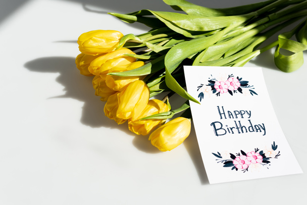 φως του ήλιου σε κίτρινες τουλίπες κοντά ευχετήρια κάρτα με χαρούμενα γράμματα γενέθλια σε λευκό - Φωτογραφία, εικόνα