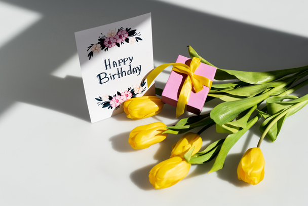sunlight on yellow tulips near present and greeting card with happy birthday lettering on white - Photo, Image