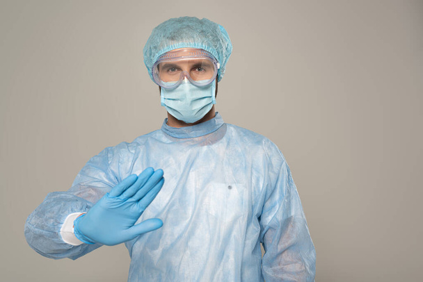 Доктор в медицинской маске показывает стоп-жест на камеру, изолированную на сером
 - Фото, изображение