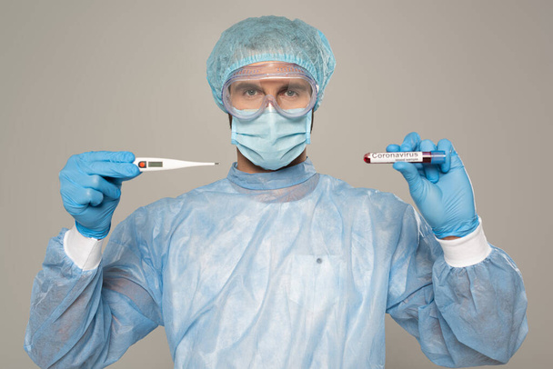 Arzt in medizinischer Maske hält Thermometer und Reagenzglas mit Blutprobe und Coronavirus-Schriftzug isoliert auf grau - Foto, Bild