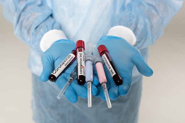 Cropped view of doctor holding test tubes with blood samples and coronavirus lettering and syringes isolated on grey - Foto, Bild