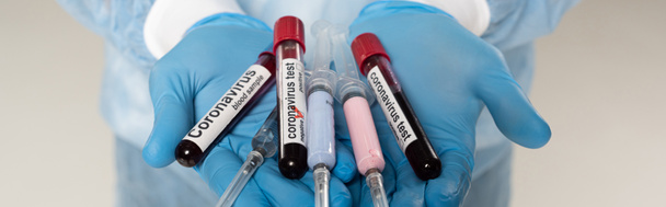 Vista recortada del médico sosteniendo tubos de ensayo con muestras de sangre y letras de coronavirus y jeringas aisladas en gris, tiro panorámico
 - Foto, Imagen