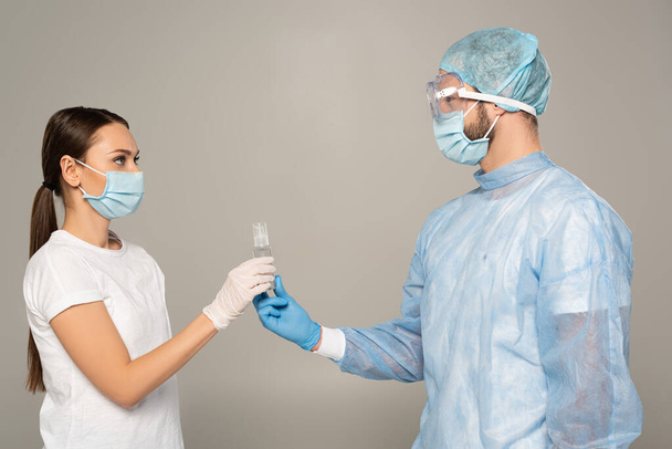Vista lateral do médico dando garrafa de desinfetante de mão para a mulher em máscara médica isolada em cinza
 - Foto, Imagem