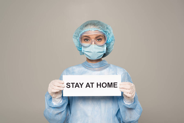 Médico en googles protectores y tarjeta de retención de máscara médica con letras de estancia en casa aislado en gris
 - Foto, Imagen