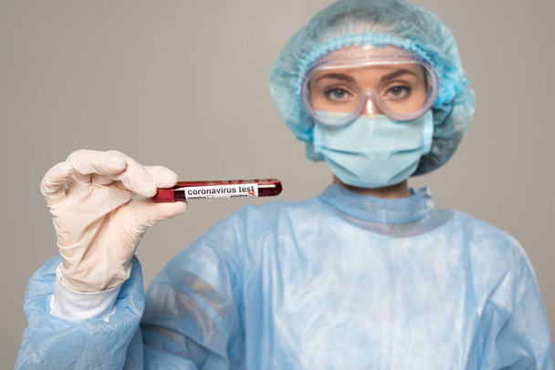 Enfoque selectivo del médico en máscara médica y tubo de prueba de retención de la tapa con muestra de sangre y letras de coronavirus aislados en gris
 - Foto, Imagen