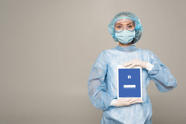 KYIV, UKRAINE - APRIL 1, 2020: Doctor in safety googles and medical mask holding digital tablet with facebook app isolated on grey - Fotografie, Obrázek