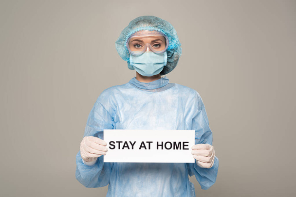 Arts in medisch masker en cap holding card met verblijf thuis belettering geïsoleerd op grijs - Foto, afbeelding
