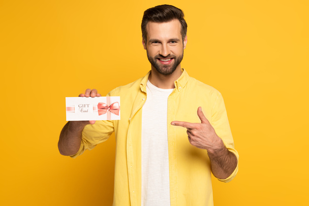 Smiling man pointing with finger at gift card on yellow background - Photo, Image