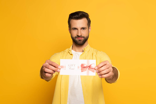 Selective focus of handsome man holding gift card and looking at camera on yellow background - Foto, afbeelding