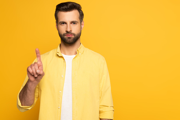 Hombre guapo apuntando hacia arriba con el dedo y mirando a la cámara en el fondo amarillo
 - Foto, Imagen