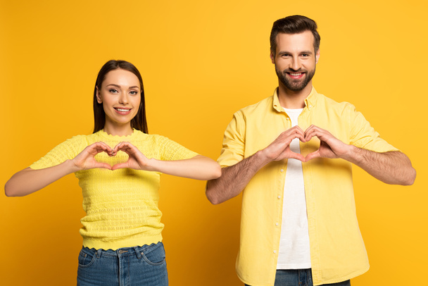 Smiling couple showing heart sign and looking at camera on yellow background - Foto, Imagem