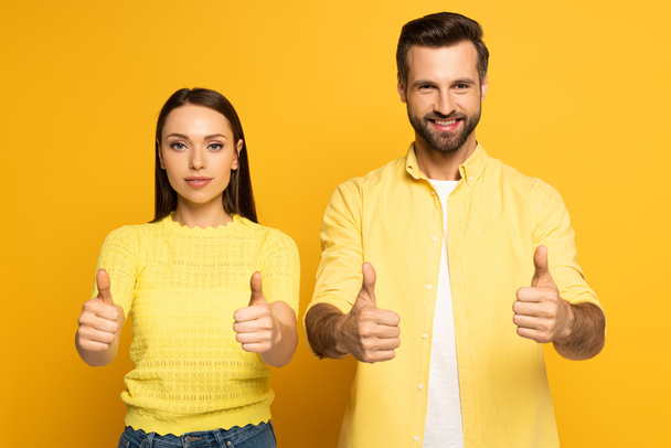Young couple showing tums up at camera on yellow background - Fotografie, Obrázek