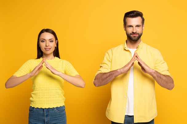 Молодая улыбающаяся пара показывает домой жест в языке жестов на желтом фоне
 - Фото, изображение