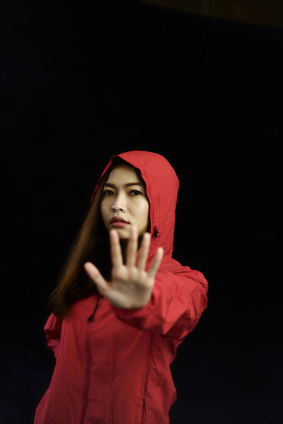 Mujer asiática que usa abrigo rojo con signo de mano "stop" sobre fondo negro, concepto de violencia (low key
) - Foto, imagen