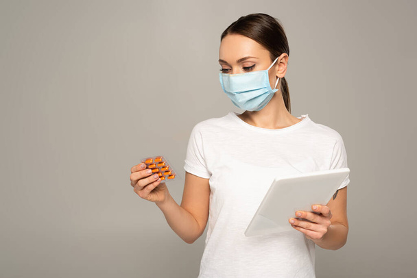 Woman in medical mask holding blister with pills and digital tablet  isolated on grey  - Φωτογραφία, εικόνα