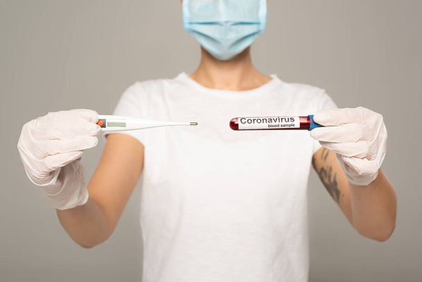 Обрізаний вид дівчини в медичній масці, що тримає термометр і пробірку з зразком крові та коронавірусом, ізольовані на сірому
  - Фото, зображення