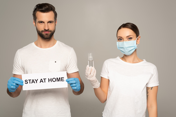 Jong echtpaar in medisch masker en latex handschoenen holding card met verblijf thuis belettering en fles van de hand sanitizer geïsoleerd op grijs  - Foto, afbeelding