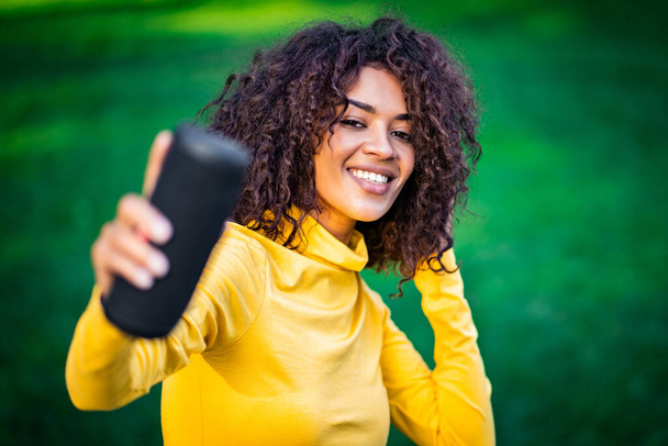 Modern trendy african american girl listening to music by wireless portable speaker.Young beautiful woman enjoying, dancing in park. - Фото, изображение